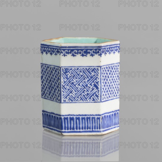 Hexagonal bowl holder in the shape of a prism, decorated with three bands of geometric borders and meander ornament. Porcelain body with white glaze outside; overglaze decoration with cobalt enamel; interior and foot glazed in turquoise; bronze foot and rim edge. Signed. Incense container, colored porcelain, Qing dynasty Qianlong style, 18th–19th century?, Height 7.5 cm, width 5.8 cm., A hexagonal ceramic vessel stands on a reflective surface against a pale gray background, showing a white body with decorative bands of deep blue patterns and a pale green interior; the top rim has a thin brown edge, the upper band features a scrolling blue border on white, the central band displays alternating intricate geometric blue motifs on white, and the lower band has vertical blue fluting on white, with a faint reflection visible beneath.