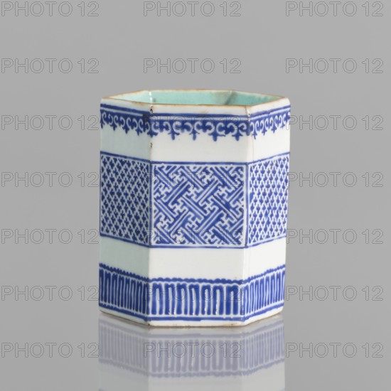 Hexagonal bowl holder in the shape of a prism, decorated with three stripes of borders with geometric ornament. Porcelain body with white glaze outside, overglaze decoration with cobalt enamel; inside and foot glazed in turquoise. Bronze foot and rim edge. Incense container, colored porcelain with meander ornament, Qianlong style Qing dynasty, 1644–1911, signed. Possibly 19th century, China. Height 7.5 cm, width 5.8 cm., A hexagonal ceramic vessel with a glossy surface sits against a neutral gray background, predominantly white with decorative bands of deep blue forming intricate geometric and repeating patterns around its sides; the upper interior shows a pale turquoise glaze, and the rim has a thin brownish edge. The blue decoration includes a scroll-like border near the top, a central band of interlocking angular motifs framed by finer textured panels, and a lower band of vertical brushstroke-style marks, with the vessel and its colored patterns reflected faintly on the smooth surfa...