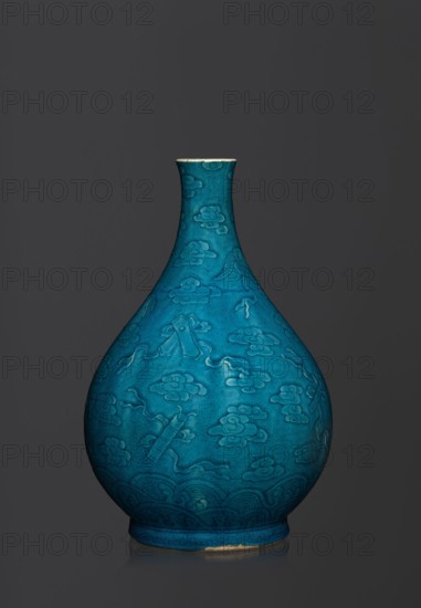 Vase in the shape of an onion-shaped bottle, Onion-shaped bottle-vase with a narrow, short neck. The vessel is decorated with a flat relief creating a composition combining a cloud and babao emblems eight precious objects or eight basic symbols. Glaze is single-color, dark, in green-blue tones. Qing 1644–1911 style; 16th–17th century . Chinese ceramics. Height 27 cm; weight 2.6 kg.; Chinese culture., A teardrop-shaped ceramic vase with a narrow neck and rounded body displayed against a dark gray background, the vase glazed in a rich turquoise-blue with a slightly crackled surface and subtle variations in tone, featuring an all-over raised pattern of stylized cloud and ribbon motifs and curved wave-like designs near the base, with a thin unglazed rim showing a light beige edge and a small exposed area of the base in a similar beige.