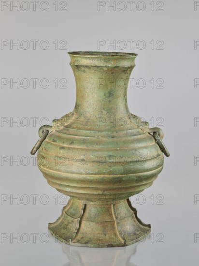 Hu-type vessel; maker: Hôun; Japanese; ceremonial bronze wine vessel hu; 1st half of the 20th century Meiji/Taishô periods; globular body with narrow, rather high neck and conical stem; decorated with profiled circles around the body; two mascarons with handles; light green artificial patina; height 40.0 cm., An aged metal vessel with a rounded body narrowing to a tall neck and flared rim, mounted on a flared segmented base; the surface shows a patina with shades of green and brown and areas of darker verdigris; the body has horizontal ridges encircling it and a pair of small looped handles on opposite sides with short hanging bars; the base has recessed panels and curved edges; the object is set against a plain light gray background and rests on a reflective surface that produces a subtle mirrored outline.