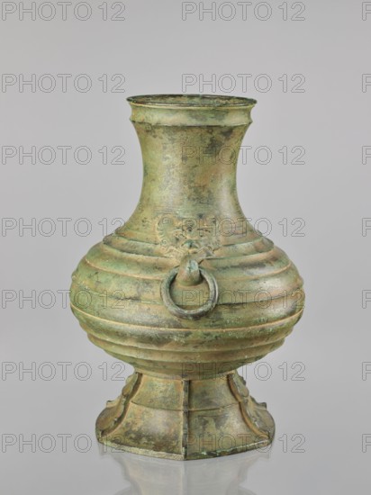 Hu-type vessel hu, bronze wine vessel, Japanese, Meiji–Taisho period, 1st half of the 20th century; globular body with narrow high neck and conical stem; decorated with profiled circles around the body; two mascarons serving as handles; light green artificial patina; height 40.0 cm., An aged metal vessel with a bulbous body, flared neck, and a pedestal base, showing a mottled green and brown patina; the body features horizontal ridges and a raised decorative face with a circular ring hanging from its mouth, and the base has flared, segmented panels with similar weathered green and brown tones against a neutral gray background.