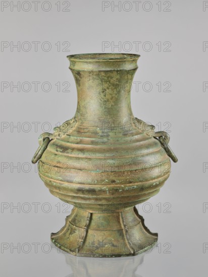 Hu-type vessel hu, Japanese, Bronze wine vessel. 1st half of the 20th century Meiji/Taisho periods, 1868–1926. Globular body with narrow, rather high neck and conical stem. Decorated with profiled circles around the body. Two mascarons serve as handles. Light green artificial patina. Height 40.0 cm. Maker role/classification: Hôun craftsman., An aged metal vessel with a rounded, ribbed body, a flaring neck, and a flared, segmented pedestal base, showing a mottled surface patina in green and brown tones; the body has decorative ringed bands and a pair of small looped handles on opposite sides with short bars through them, and the surface displays areas of darker brown, verdigris green, and lighter green speckling against a neutral gray background.