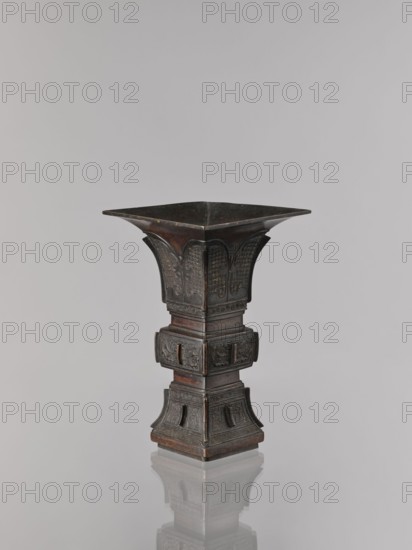Fangzun vase, Chinese art, Chinese culture, plant ornament, banana plants, meander ornament, taotie iconography, ceremonial/ritual vessel, Qing dynasty 1644–1911, Qing style, 19th century, China place of issue?, vessel, height 21.4 cm, width 13.3 cm, A tall, ornate vessel with a flared square top narrowing into a sculpted midsection and widening again at a decorative square base, finished in dark bronze tones with areas of brownish patina; the surface shows intricate carved relief panels and raised decorative motifs on each face, with curved flared panels near the top and rectangular recessed panels near the base, and the object sits on a reflective surface that produces a faint mirror image below.