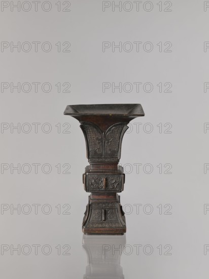 Fangzun vase, Chinese art, Chinese culture, plant ornament, banana plants, meander ornament, taotie iconography, ceremonial vessel, ritual vessel, Qing dynasty 1644–1911, Qing style, 19th century, China place of issue ?, vessel; height 21.4 cm, width 13.3 cm, An upright, dark brown and black carved object with a flared square top and a stepped square base, showing ornate carved panels and repeating geometric and curving motifs on its sides, standing on a reflective pale gray surface against a light gray background, the object casting a soft reflection below it.