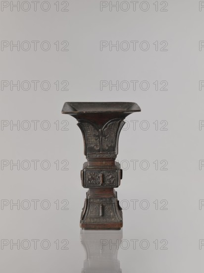 Fangzun vase, Chinese art, Chinese culture, ornament: plant banana plants, meander, iconography: taotie, ceremonial/ritual vessel, Qing dynasty 1644–1911, Qing style, 19th century, China place of issue ?, vessel, height 21.4 cm, width 13.3 cm, A single tall, ornate vessel stands centered against a pale gray background; it is dark brown with areas of reddish-brown patina and subtle black accents, showing a squared, flared rim at the top, a narrow midsection with carved rectangular panels, and a broader stepped base with decorative relief patterns, and the object casts a faint reflection on the smooth surface beneath it.