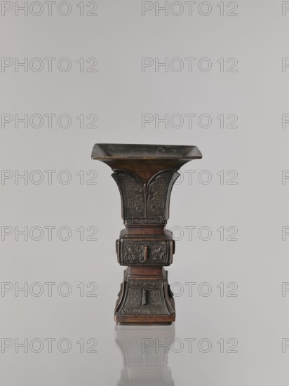 Fangzun vase, Chinese, Qing dynasty 1644–1911, 19th century, ceramic vessel; ritual/ceremonial vessel featuring plant ornament banana plants, meander ornament, taotie iconography; height 21.4 cm, width 13.3 cm., A single ornate object stands centered against a pale gray background, appearing to be made of dark brown and black metal with areas of warm brown patina; the object has a flared top with a shallow square bowl, a tapered neck decorated with carved patterns, a rectangular central section with raised relief details, and a broad square base adorned with geometric and floral motifs; the surface shows polished highlights and darker recessed areas, and the object casts a faint reflection on the glossy surface below.