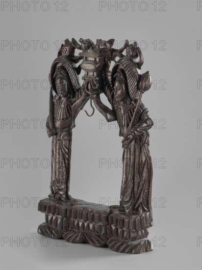 Stand, Chinese, Chinese art, figurative motifs, women, Qing 1644–1911, stand for hanging a gong, 19th century, China, varia; height 45.5 cm, width 29.5 cm, A dark brown carved wooden sculpture of two standing figures facing each other on a shared carved base, each figure wearing ornate headdresses and long patterned robes, with detailed facial features and folded hands; they support an elaborately carved central element between them that includes stylized animal heads and decorative motifs, and one figure holds a long staff topped with a rounded form; the base and figures show carved geometric and floral patterns and a polished surface in shades of dark brown with lighter brown highlights, set against a plain light gray background.