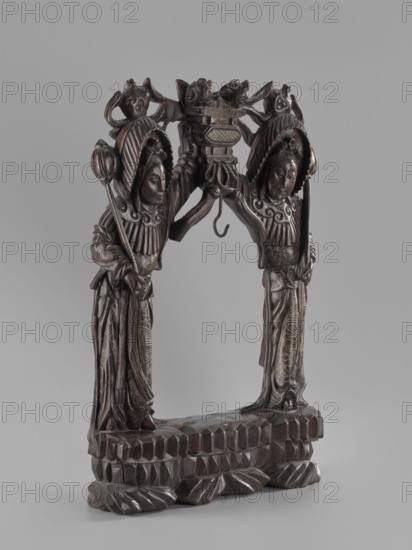 Stand, Chinese, Chinese art, figurative motifs, women, Qing 1644–1911, Stand for hanging a gong, 19th century, China, varia; height 45.5 cm, width 29.5 cm, A carved wooden sculpture depicting a pair of robed figures standing back to back and facing inward toward a central arch formed by their raised arms, each figure wearing ornate headgear and holding a staff, with elaborate carved patterns on their garments and detailed facial features, a carved decorative element with animal-like faces at the top center where the arms meet, a hooked piece suspended beneath the arch, and a textured stepped base with faceted and leaf-like forms; visible colors include dark brown wood tones and subtle lighter brown highlights against a neutral gray background.