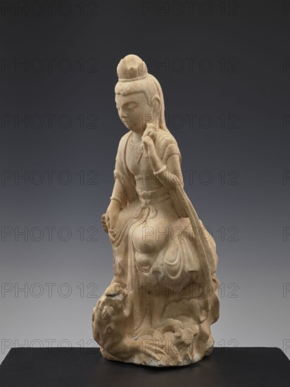 Guanyin with qilin, Chinese, Qing 1644–1911, goddess of Chinese Buddhism with unicorn qilin iconography, 19th century, China, sculpture figurine, height 47.5 cm, A single pale beige stone statue of a seated figure with a rounded topknot and long hair draped over the shoulders sits on a dark flat base; the figure wears flowing robes with carved folds and decorative elements, one hand raised near the chest holding a small rounded object and the other hand resting near the knee, while a carved creature with a rounded face and visible paws is positioned at the figure’s feet. The background is a smooth gradient from dark gray at the top to lighter gray toward the bottom, and the visible colors are beige, dark gray, and light gray.