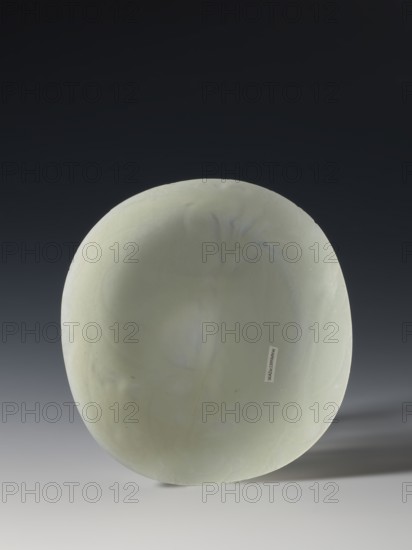 Plaque-lid, 19th century Qing dynasty 1644–1911, China; bas-relief carved jade with engraving; decoration of bats and plant motifs; three double recesses on the bottom; height 14.2 cm, width 15.4 cm., A smooth, rounded object with a pale green surface sits against a dark to light gradient background, showing subtle variations in green and faint bluish-gray veining; the object casts a soft shadow to the right and rests on a light gray surface, and the overall scene includes shades of dark gray, black, pale green, bluish-gray, and light gray.