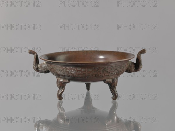 Ding vessel, Ding vessel with three legs and two handles; used for ritual hand washing. Bronze. 19th century late Edo–Meiji, 1603–1912. Japan. Height 9.1 cm; width 24.2 cm; diameter 19.6 cm. Japanese tripodal vessel., A low round metal bowl with a brown surface sits on a reflective gray surface against a light gray background, the bowl supported by three curved legs shaped like stylized animal forms and flanked by a pair of short upward-curving handles, the exterior rim and body showing carved geometric patterns and the interior appearing smoother and slightly darker, with visible reflections of the bowl and legs on the surface below, and colors including shades of brown, bronze, gray, and muted black.