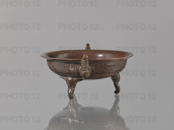 Ding vessel, Tripod bronze vessel with three legs and two handles; used for ritual hand washing. Japanese, Edo–Meiji periods, 19th century 1603–1912. Height 9.1 cm; width 24.2 cm; diameter 19.6 cm., A small round bronze bowl with a reddish-brown patina sits on a reflective surface, supported by a group of three short legs shaped like stylized animal heads and curved appendages; the bowl's exterior is decorated with a band of low-relief geometric patterns and carved motifs, and a few raised looped handles project from the rim. The visible colors are reddish-brown, dark brown, and hints of greenish-brown on the metal, with a neutral gray background and softer gray reflections on the surface beneath the bowl.