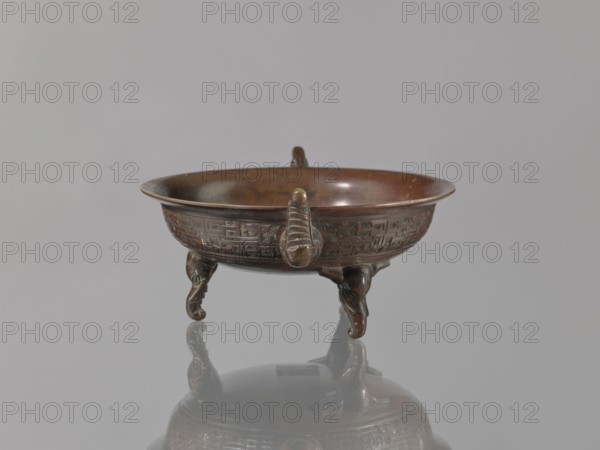 Ding vessel, gift, Japanese art, Japanese culture, tripod, Edo 1603–1868, bronze, Meiji 1868–1912. Ding vessel with three legs and two handles. Used for ritual hand washing. 19th century Edo/Meiji, Japan. Vessel; height 9.1 cm, width 24.2 cm, diameter 19.6 cm., A shallow round bowl with ornate relief decoration sits on three sculpted legs, each leg curving and ending in a rounded foot, and the bowl has a small raised handle-like projection on its rim; the object shows a metallic surface with brown and bronze tones and areas of darker patina, and it is reflected on a smooth gray surface beneath; the background is a uniform light gray.