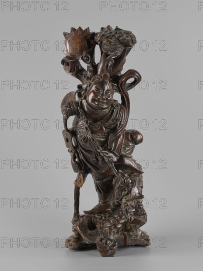 Mythical figure, unknown sculptor; Chinese and Japanese art; pine trees; mythology; beasts fantasy; unicorns iconography; Qing 1644–1911; fantasy animals; 2nd half of the 19th century; China place of origin?; Japan place of origin?; sculpture, figurine; height 47.0 cm, A carved wooden figure with a dark brown polished surface depicting a smiling person in flowing robes standing on an irregular base, holding a staff in one hand and a chain or rope in the other, with detailed carved features including a round face, stylized hair or topknot, layered clothing, and ornamental elements; behind the figure rises a carved tree-like form with textured foliage and a circular sun-like shape, and the sculpture shows areas of lighter brown where the wood grain and wear are visible against a neutral light gray background.