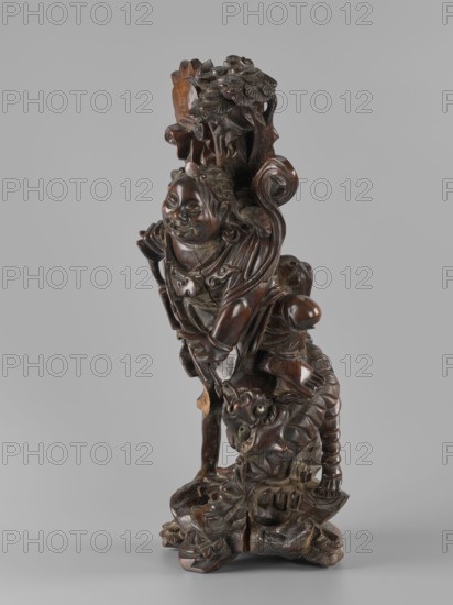 Mythical figure, unknown sculptor, Chinese and Japanese art; pine trees; mythology; fantasy beasts; unicorns iconography; Qing dynasty 1644–1911; fantasy animals; Japanese culture; 2nd half of the 19th century; China place of origin ?; Japan place of origin ?; sculpture, figurine; height 47.0 cm, A carved wooden sculpture depicting a robed figure with a rounded smiling face and stylized hair, standing on a textured base that includes a coiled, scaled creature beneath the figure's feet; the carving shows flowing drapery, curling ornamental elements, and a cluster of layered foliage near the top. The visible colors are dark brown wood tones with lighter brown highlights and small areas of muted greenish patina.