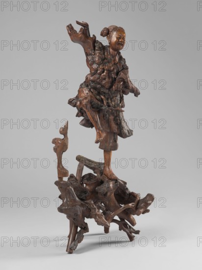 Dancing with a bird, unknown sculptor, figurative motifs of birds and cranes, dance iconography; Asian — Chinese; 19th century; sculpture; height 77 cm, A carved wooden figure stands on a gnarled wooden base, showing a person with an uplifted arm and a playful facial expression, wearing a textured garment with flowing folds and layered details; the carving includes rough, knotty wood surfaces and smoother polished areas, and a small carved animal-like form is attached near the base. The visible colors are brown tones in the wood, darker brown in the knotted areas, and a neutral gray background.