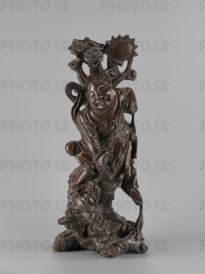 Mythical figure, unknown sculptor; Chinese/Japanese art; pines; fantastical beasts; fantasy animals; mythology; unicorns iconography; Qing dynasty 1644–1911; 2nd half of the 19th century; sculpture, figurine; height 46 cm; possible China or Japan origin., A carved wooden sculpture depicting a robed figure with a rounded, smiling face standing on a textured base that includes a stylized creature head; the figure wears flowing garments with carved folds and holds a staff or branch that extends upward into a leafy cluster and a radiating circular form, while the carving shows detailed patterns, curved elements, and smooth polished surfaces in varying shades of brown against a pale gray background.