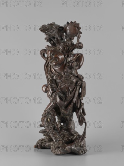 Mythical figure, unknown sculptor, Chinese and Japanese art and culture influences, pines, fantastical beasts, fantasy animals, mythology, unicorns iconography, Qing dynasty 1644–1911, 2nd half of the 19th century, possibly China or Japan, sculpture figurine, height 46 cm., A carved wooden figure in dark brown with polished highlights stands against a plain gray background, showing a robed person with a smiling face and closed eyes, ornate flowing garments, and detailed braided or beaded adornments; the figure holds or leans against a stylized staff or tree-like form topped by layered floral or leaf motifs and a sunburst-like disk, while the lower portion rests on a carved base featuring swirling forms and a dragon- or animal-like head with textured scales and curled elements in similar dark brown tones.