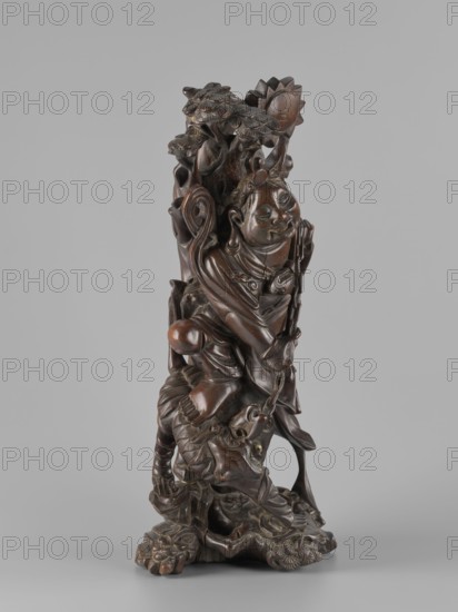 Mythical figure; unknown sculptor; Chinese art; Chinese culture; pines; fantastical beasts; Qing dynasty 1644–1911; Asia; fantasy animals; mythology; unicorns iconography; Japanese art; Japanese culture; 2nd half of the 19th century; China place of origin?; Japan place of origin?; sculpture; figurine; height 46 cm, A carved wooden figure displayed against a plain light gray background, showing a vertical sculptural composition with smooth dark brown and reddish-brown tones. The carving depicts a robed figure with a rounded face and detailed facial features, wearing layered garments and ornate decorative elements, with a few flowing ribbon-like forms around the shoulders. Above and behind the figure are carved tree-like forms and a stylized floral or sunburst motif. The lower portion shows a textured base with carved leaves and organic shapes and a creature with a rounded snout and pronounced eyes integrated into the composition. The surface varies between polished smooth areas and more...