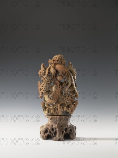 Phoenix with a dragon and a pearl on a base; creator, Chinese; Qing dynasty 1644–1911, 18th century; sculpture/figurine in the round, height 36.0 cm, width 17.4 cm; materials: likely carved figurines, depicts phoenix and dragon iconography with a pearl, clouds, and rocks., An intricately carved ornamental object stands on a carved openwork base, the piece showing a group of organic, flowing forms resembling stylized birds and flora. The carving displays a range of warm beige, tan, and brown tones with subtle variations and some darker mottled areas, while the base has deeper brown and grayish-brown coloration with pitted and porous textures. The top portion features layered, lacy carving with curved feather-like and leaf-like elements, rounded protrusions, and hollowed openings that create interplay of light and shadow. The overall silhouette is vertical and tapering, with delicate carved extensions that curl and overlap, and a smooth, lighter lower surface beneath the base where the o...