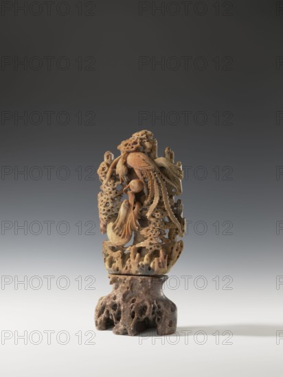 Phoenix with a dragon and a pearl on a base; unknown creator; Chinese; Qing dynasty 1644–1911; 18th century; sculpture/figurine; materials: pearls decorative, depicts dragon with a pearl, phoenix, clouds, rocks; height 36.0 cm, width 17.4 cm., The image shows an intricately carved vertical sculpture in warm beige and brown tones standing on a darker brown, porous base; the carved section features flowing organic forms including curved, featherlike elements and rounded, layered shapes with many small perforations and textured surfaces, creating a sense of depth and interlocking pieces, and the background is a smooth gradient from dark gray at the top to light gray near the bottom.