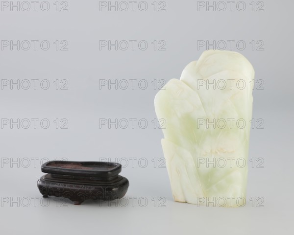 Small screen on a base, Chinese, Qing dynasty 1644–1911. Small screen with an oval cross-section made of pale green jadeite. Engraved decoration: composition with pine, grass, lingzhi mushrooms on rocks; at the top a crescent with the inscription yue. Wooden base, elongated, tapering on the shorter side, on four short legs one broken off. Bulging side inlaid with silver wire. 18th–19th century. Sculpture; screen. Dimensions: height 14.3 cm, width 9.1 cm; base height 8.7 cm, width 4.7 cm., A pale green and white carved stone object with smooth rounded tops stands upright on the right, showing subtle veining and lighter patches of cream and very light yellow; on the left sits a dark brown, almost black low wooden stand with a shallow rectangular inset and decorative carved patterns on its sides, with hints of reddish brown in the inset and on small feet, all set against a soft light gray background.