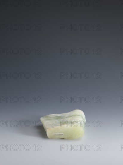 Small screen on a base, Chinese, Qing 1644–1911. Small screen with an oval cross-section made of pale green jadeite. Engraved decoration: composition with pine, grass, lingzhi mushrooms on rocks; at the top a crescent with the inscription yue. Wooden base, elongated, tapering on the shorter side, on four short legs one broken off. Bulging side inlaid with silver wire. 18th–19th century, China. Sculpture; screen. Dimensions: screen height 14.3 cm, width 9.1 cm; base height 8.7 cm, width 4.7 cm., A small irregularly shaped object rests on a smooth surface, appearing to be made of a translucent pale green material with areas of slightly darker green and hints of white; the object has smooth rounded edges and subtle carved or molded flowing patterns on its top, with a faint shadow cast to one side against a gradient background that transitions from dark gray at the top to lighter gray toward the bottom.