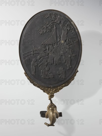 Framed oval screen, Chinese, Qing 1644–1911, depicting children and children's games. Oval screen made of black slate in a gilded bronze frame topped with a frog figurine. 19th–20th century, China; sculpture/screen; height 42.0 cm., An oval handheld mirror with a dark, carved front showing a scene of people beneath a tree and a pavilion, mounted in an ornate gold-colored frame with a decorative gold-colored handle shaped like a stylized fish; visible colors include dark gray or black on the carved surface and gold tones on the frame and handle, with a pale neutral background.