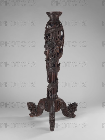 Pedestal, Chinese, Qing 1644–1911, 17th–19th century. Sculpture, height 128 cm. Portrays a dragon winding upwards around the shaft with fantastical beasts and dragons iconography., A tall, intricately carved dark brown wooden stand with a slender, columnar body wrapped by a carved dragon motif and flowing, flame-like carved details; the base spreads into three carved legs ending in stylized dragon heads and scaled tails, all in dark brown wood against a neutral light gray background.
