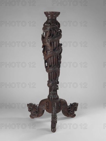 Pedestal, unknown sculptor; Chinese; Qing dynasty 1644–1911; sculpture, 17th–19th century; height 128 cm. Chinese art; dragons and fantastic beasts iconography; dragon winding upwards around the shaft., A tall, vertical carved wooden stand with a dark brown finish, featuring a spiraling carved dragon motif wrapping around the central column; the column is composed of fluted vertical elements and carved scales, with intricate openwork and layered carved details. The base has three carved legs ending in stylized dragon or animal heads and textured scale patterns, and the top flares into a carved circular rim with decorative carving. The visible colors are dark brown wood tones against a light gray background.