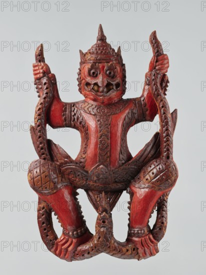 Demon with snakes - wall sculpture, unknown sculptor, gift; snakes; art of Southeast Asia; Southeast Asia culture; religion; 19th century; Indonesia; sculpture; height 44 cm, A carved wooden figure painted predominantly red with areas of brown showing through, depicting a stylized humanoid with a pointed headdress, large round eyes, a broad nose, and an open mouth showing carved teeth; the figure has a textured torso with raised decorative patterns, bent arms raised to the sides holding sinuous carved elements, and bent legs with patterned knee guards and clawed feet; the carving includes pierced and scalloped details along the limbs and the sinuous elements, and there is a neutral pale gray background.