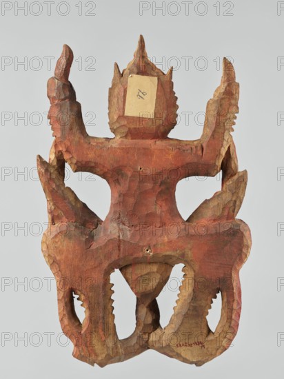 Demon with snakes - wall sculpture, unknown sculptor, snakes, art of Southeast Asia, Southeast Asia culture, religion, 19th century, Indonesia, sculpture, height 44 cm, A carved wooden object with a rough, faceted surface and an overall reddish-brown color, showing areas of lighter tan wood where the surface is worn or unpainted. The carving has a symmetrical form with two upward-pointing horn-like projections at the top, a central pointed crest, and curved arching elements on either side that create open spaces. The lower portion features rounded, scalloped cutouts with jagged inner edges and a central conical protrusion that tapers downward. The wood shows tool marks and uneven texture throughout, with some darker brown shadows in recessed areas and subtle variations of red, brown, and tan across the piece. The background is a neutral light gray.