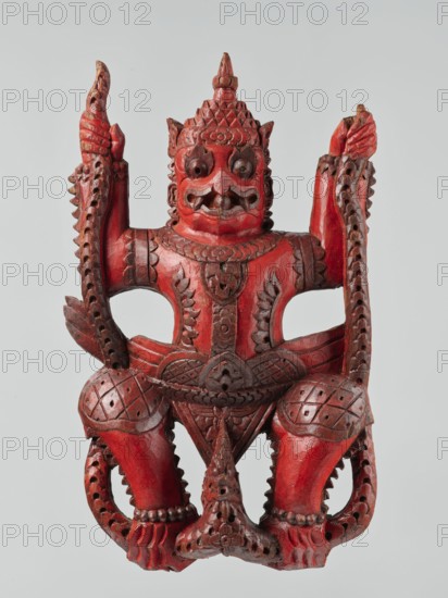 Demon with snakes - wall sculpture, unknown sculptor, snakes, Southeast Asia, Southeast Asian art, demons, religion, 19th century, Indonesia, sculpture, height 44.0 cm, width 27.0 cm, A carved wooden figure painted primarily red with areas of darker brownish-red in the carved recesses, depicting a stylized humanoid creature with a fierce facial expression, wide round eyes, prominent brows, flared nostrils, an open mouth with visible teeth, and a pointed headdress with geometric patterns; the figure has raised arms holding long carved elements with pierced decorative holes, ornate chest and waist ornamentation with carved floral and geometric motifs, detailed knee guards with crosshatched patterns, muscular legs with beaded anklets, and curved tail-like or decorative extensions along the sides, all shown against a pale gray background.