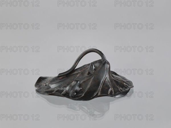 Lotus Leaf with Two Frogs, Ryûbundô,  craftsman author; Japanese; frogs; paperweight; lotus leaf; sculpture; Japan, 2nd half of the 19th century; height 6.7 cm, width 18.2 cm., A dark metallic object shaped like a large, slightly crumpled leaf with raised veins and organic folds sits on a smooth reflective surface, showing a muted reflection beneath it; a curved handle arches from one side to the other, and the surface of the object displays subtle highlights and worn areas in dark brown and black tones against a pale gray background.
