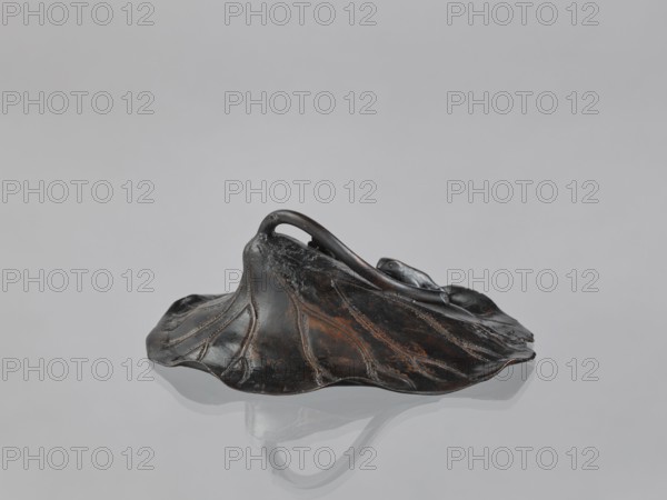 Lotus Leaf with Two Frogs, Ryûbundô,  craftsman author; Japanese; frogs; paperweights; lotuses; leaves; sculpture; Japan, 2nd half of the 19th century; height 6.7 cm, width 18.2 cm, A dark, irregularly shaped sculptural form resembling a wilted leaf or hat sits on a smooth, reflective surface; the object shows a mix of black and brown tones with subtle reddish-brown highlights and worn areas, and it has raised, vein-like ridges and a curved handle-like loop rising from its center, with a soft reflection of the object visible on the pale gray surface beneath.