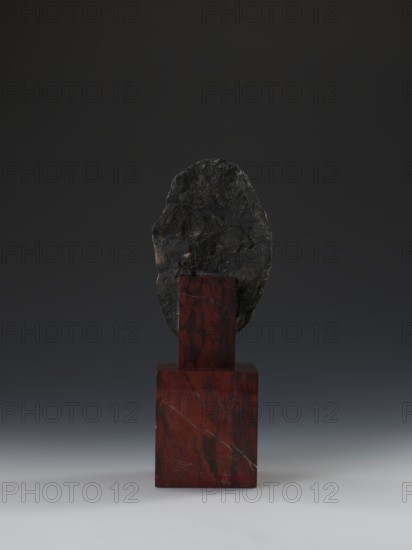 Head of Guanyin Bodhisattva, Chinese, Guanyin iconography, Qing style, polychrome-coated feldspar with cube-shaped base of dark red marble, 19th century Qing dynasty, 1644–1911, China, sculpture, height 26.0 cm, A dark gray, irregularly shaped rock is mounted on a polished red stone pedestal with visible darker veins; the pedestal consists of a larger red block topped by a smaller red column, both showing marbled patterns. The background is a smooth gradient from deep charcoal at the top to a lighter gray near the bottom, and the surface beneath the pedestal is a soft gray transitioning to an off-white foreground. The visible colors are dark gray, black, red, maroon, and shades of gray.