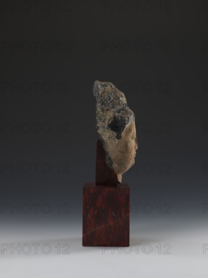 Head of Guanyin Bodhisattva, unknown sculptor; polychrome-coated feldspar fragment of a statue with cube-shaped base made of dark red marble; Chinese; Guanyin iconography; Qing dynasty 1644–1911; 19th century; sculpture; height 26.0 cm; goddesses., A weathered stone fragment resembling a carved face is mounted on a rectangular dark reddish-brown wooden base; the stone shows mottled tones of beige, tan, gray, dark gray, and hints of green, with rough textured areas and smoother carved facial features visible in profile, set against a smooth gradient background that transitions from dark gray at the top to lighter gray near the bottom.