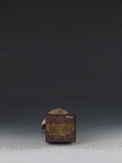 Head of Guanyin Bodhisattva, Chinese, Guanyin iconography, Qing style, goddesses. Fragment of a statue made of polychrome-coated feldspar with cube-shaped base of dark red marble. 19th century, China. Sculpture; height 26.0 cm., A small worn rectangular box with a distressed red exterior sits near the bottom center of the image, showing a weathered brown patch on its front and rough textured edges; behind and partially above the box is a pale rounded object with beige and light tan tones and a small dark area on one side; the background transitions from a dark charcoal at the top to a mid gray surrounding the objects and a lighter gray foreground.