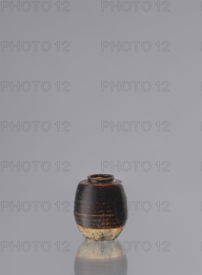 Dried tea container chaire, ; ceramics; chaire vessel; small, barrel-shaped, wheel-thrown grey clay pottery bowl with brown glaze showing rusty and brown shadows; lower unglazed part grey-brown; unsigned; 19th century; Seto, Owari, Japan; vessel/container; height 8 cm, dia. 6.2 cm; used for tea., A small rounded jar sits centered at the bottom of the image against a plain light gray background, showing a glossy dark brown to almost black upper body with subtle reddish highlights and a worn lighter beige base; the jar has a narrow rim at the top and a smooth reflective surface that creates a faint mirrored reflection beneath it.