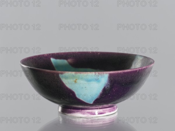 Bowl, ; vessel, ceramic, decorative; Qing dynasty 1644–1911, 18th–19th century; Chinese; purple glaze with celadon spots; manganese and copper, glaze; height 6.7 cm, diameter 18.1 cm., A shallow round bowl with a low foot, shown against a neutral gray background, has a predominantly deep purple exterior and interior with irregular patches of bright turquoise-blue and hints of turquoise-green. The rim shows variations of purple and lighter mauve, and the foot has bands of purple and pale off-white. The surface displays subtle gloss and tiny speckled textures, and a soft reflection of the bowl appears on the flat surface beneath it.