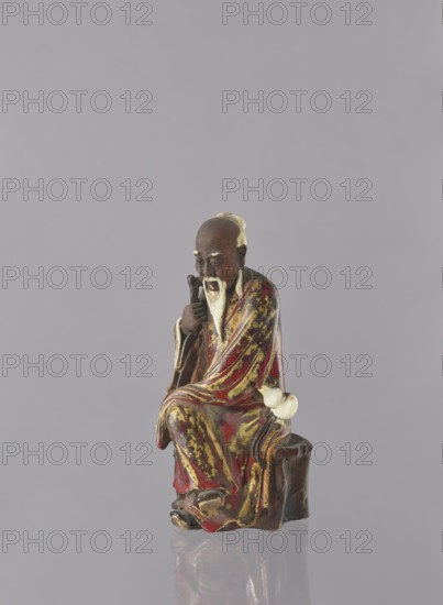 Chinese Taoist immortal Jap. sennin, Japanese art, Japanese culture, Immortals iconography, figurine, ceramic, male figurine, multi-colored glaze: white, red, brown, yellow, 19th century, Japan, possibly Seto, height 21.6 cm, A small seated figurine of an elderly man with a long white beard and mustache, wearing a flowing robe that shows a mix of red, gold, and dark brown colors, seated on a dark brown block-like stool, with one hand resting on his knee and the other holding a staff or cane, and a pale off-white gourd-shaped object tucked at his side; the background is a plain light gray and the surface beneath the figurine is reflective, showing a faint mirrored reflection.