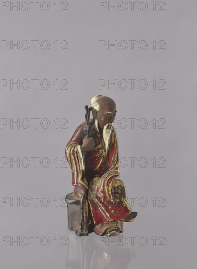 Chinese Taoist immortal Jap. sennin, Japanese art, Japanese culture, Immortals iconography, figurine, ceramic, male figurine, multi-colored glaze: white, red, brown, yellow, 19th century, Japan, possibly Seto, height 21.6 cm, A small seated figurine of an elderly man with a long beard wearing flowing robes in red with gold accents, holding a dark staff and sitting on a small block; the figure has a brownish head with pale areas on the beard and hair, and the base and staff are dark brown, all set against a plain pale gray background.