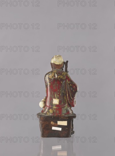 Chinese Taoist immortal Jap. sennin, Japanese art, Immortals iconography, ceramic figurine of a male immortal, multi-colored glaze: white, red, brown, yellow, 19th century, Japan, possibly Seto, height 21.6 cm, The image shows the rear view of a small figurine seated on a rectangular base against a plain light gray background. The figurine has a rounded head with a pale yellowish top and darker brown sides, and wears a robe that is predominantly deep red with patches and streaks of green and yellowish-gold. One arm rests at the figure's side holding a pale, ball-like object, and the opposite side shows a raised sleeve with darker brown inside folds. The rectangular base is dark brown with a slightly glossy surface and shows small pale beige areas where labels or stickers appear to be attached. The overall color palette includes light gray, pale yellow, brown, deep red, green, yellowish-gold, and beige.