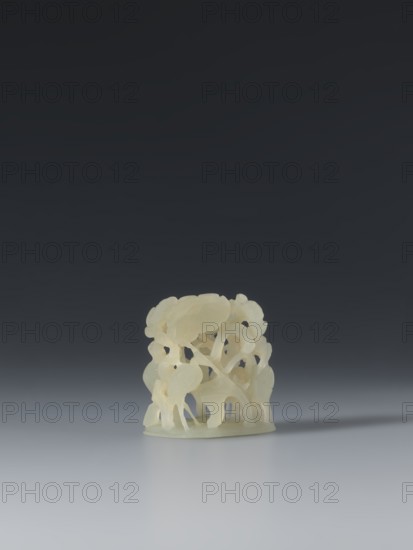 Ornament, early 17th century, China; jade; height 3.65 cm, width 3.2 cm, depth 2.5 cm. Ming 1368–1644 style. The ornament is a complex three-dimensional scene of a lotus pond: four cranes standing in natural poses among densely growing lotuses. The design was based on a thirteenth-century Yuan-era ornament. Though Mongolian nobility once wore wide-brimmed hats with jade bosses on top, by the Ming this headgear was abandoned; jade ornaments remained in use and were also applied to vessels as decorative elements for their material and craft value. Categories/subjects: bosses, lotuses, headgear, cranes, ornaments, clothing, varia., A small carved object sits on a smooth surface, depicted in pale creamy white and off-white tones; the background transitions from dark charcoal at the top to mid-gray near the surface, creating a gradient of deep gray to lighter gray; the carved piece shows a dense, intricate lattice of organic shapes with rounded, leaf-like and curved forms and hollowed openi...