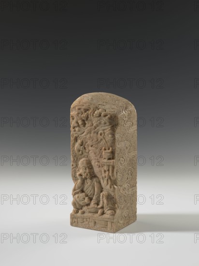 Seal stamp, lion iconography, bats iconography, arhat iconography, Chinese art, seal stamps, Chinese culture, Qing 1644–1911, bas-relief decoration, 19th century, China, seal stamp, height 23.6 cm, width 8.9 cm, depth 9.0 cm, A carved rectangular stone object with a rounded top stands on a flat surface against a gradient background. The object is a beige to light brown color with subtle darker brown variations and textured surface details. The front face features a carved human figure seated in a contemplative pose, surrounded by swirling cloud-like motifs and additional carved elements that project from the surface. The sides and top of the object display carved patterns and textured areas consistent with weathered stone. The lighting casts a soft shadow to the right and the overall scene includes shades of gray in the background and white on the surface beneath the object.