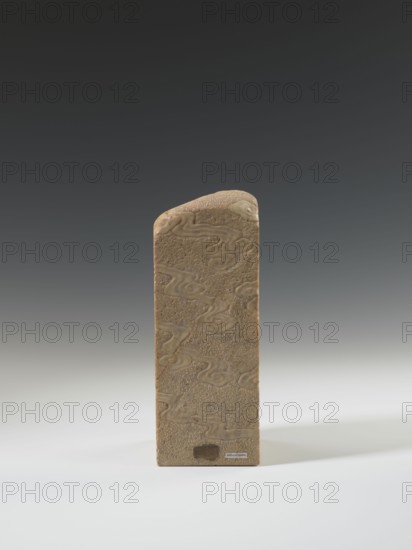 Seal stamp, unknown sculptor; lion iconography; bats iconography; arhat iconography; Chinese art; seal stamps; Chinese culture; Qing 1644–1911; bas-relief decoration; 19th century ; China place of origin; seal stamp; height 23.6 cm, width 8.9 cm, depth 9.0 cm., A tall rectangular stone object stands upright on a smooth surface, featuring a rounded top and a textured surface with flowing, cloud-like relief patterns; the stone is a warm beige to light brown color with slightly darker carved lines, set against a gradient background that transitions from dark gray at the top to light gray near the bottom, and the surface it rests on is white to pale gray.