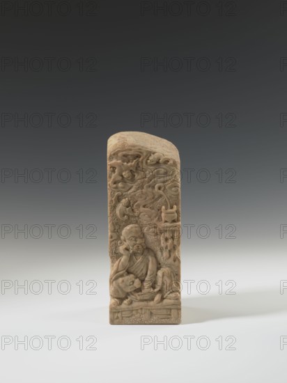 Seal stamp, unknown sculptor, lion iconography, Krieg doctor, bats iconography, arhat iconography, Chinese art, seal stamps, Chinese culture, Qing 1644–1911, Bas-relief decoration, 19th century, China, seal stamp, height 23.6 cm, width 8.9 cm, depth 9.0 cm, A carved vertical stone or ivory object in beige and light brown tones stands upright against a gradient gray background; the carving shows a seated robed figure with a contemplative expression, a rounded head, and folded garments, beside a reclining animal-like form and surrounded by flowing cloud-like motifs and decorative elements, with the base showing a patterned platform.