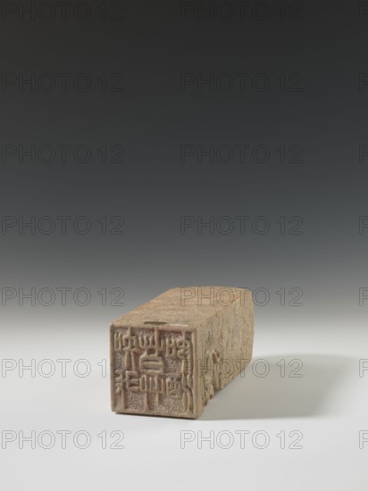 Seal stamp, lion iconography, bats iconography, arhat iconography, Chinese art, seal stamp, Chinese culture, Qing 1644–1911, bas-relief decoration, 19th century, China, seal stamp, height 23.6 cm, width 8.9 cm, depth 9.0 cm, A rectangular carved object rests on a smooth surface, shown from a low front-left angle; the object has a worn beige and light brown stone appearance with intricate carved patterns along its top and side faces and a detailed relief design on its front face, and the scene includes soft shadows beneath the object and a gradient background shifting from dark gray at the top to light gray near the surface.