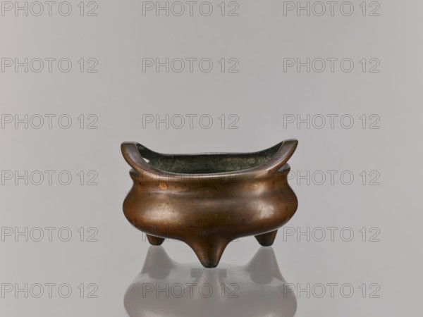 Li-type incense burner, Chinese, Li-type ritual vessel, ceremonial vessel, Ming style and Qing style influences, Xuande 1426–1435 attribution/backdated signature reading 1431, 18th century, backdated signatures, ritual use, China, height 8.8 cm, diameter 14.2 cm, A small rounded metal vessel with a warm brown and bronze patina sits on a reflective surface; it has a flared rim that curves upward at opposite sides, a dark interior, a bulbous body with subtle surface marks and spots, and three short tapered legs creating a reflected silhouette below. The visible colors include brown, bronze, dark greenish-black inside, and muted gray from the background and reflection.