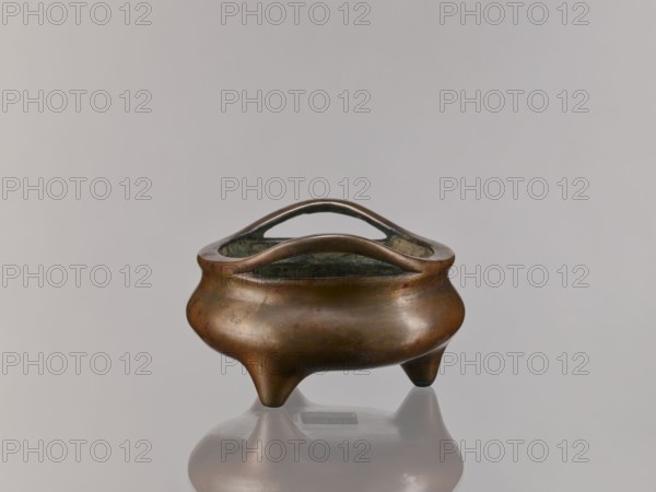 Li-type incense burner, Chinese, Li-type ritual/ceremonial vessel, Ming style Xuande, 1426–1435 with backdated signature reading 1431, Qing style Qing dynasty 1644–1911, 18th century, Chinese porcelain vessel, height 8.8 cm, diameter 14.2 cm., A small rounded metal container with a smooth surface and a warm brown patina sits on a reflective surface, showing a soft reflection beneath it; the container stands on a group of three short tapered legs and has an open top with an integrated curved handle that arches across the opening, revealing a darker interior; the background is a uniform pale gray, and the visible colors include brown, dark greenish-gray inside the vessel, and pale gray in the background and reflection.
